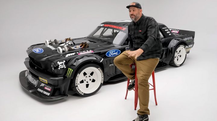 Video screenshot of Brian Scotto discussing the 1965 Ford RTR Hoonicorn V2, which is parked in the background of the shot. 